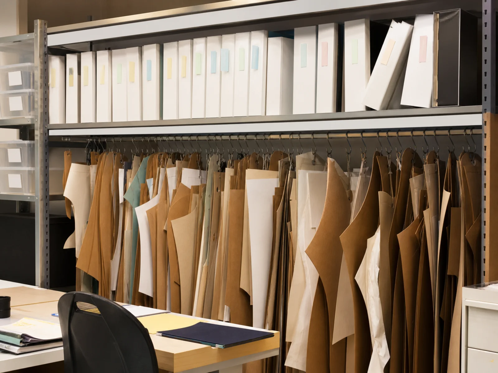 Metal shelving of blank-labelled archive boxes above a rail hung with kraft-paper pattern pieces
